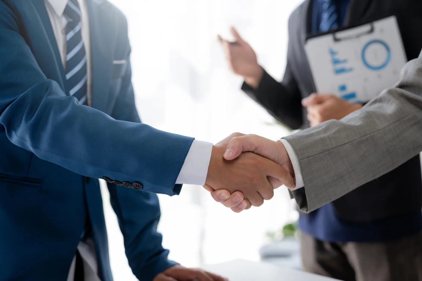 Two people shaking hands across a desk while a third holds a clipboard of charts.