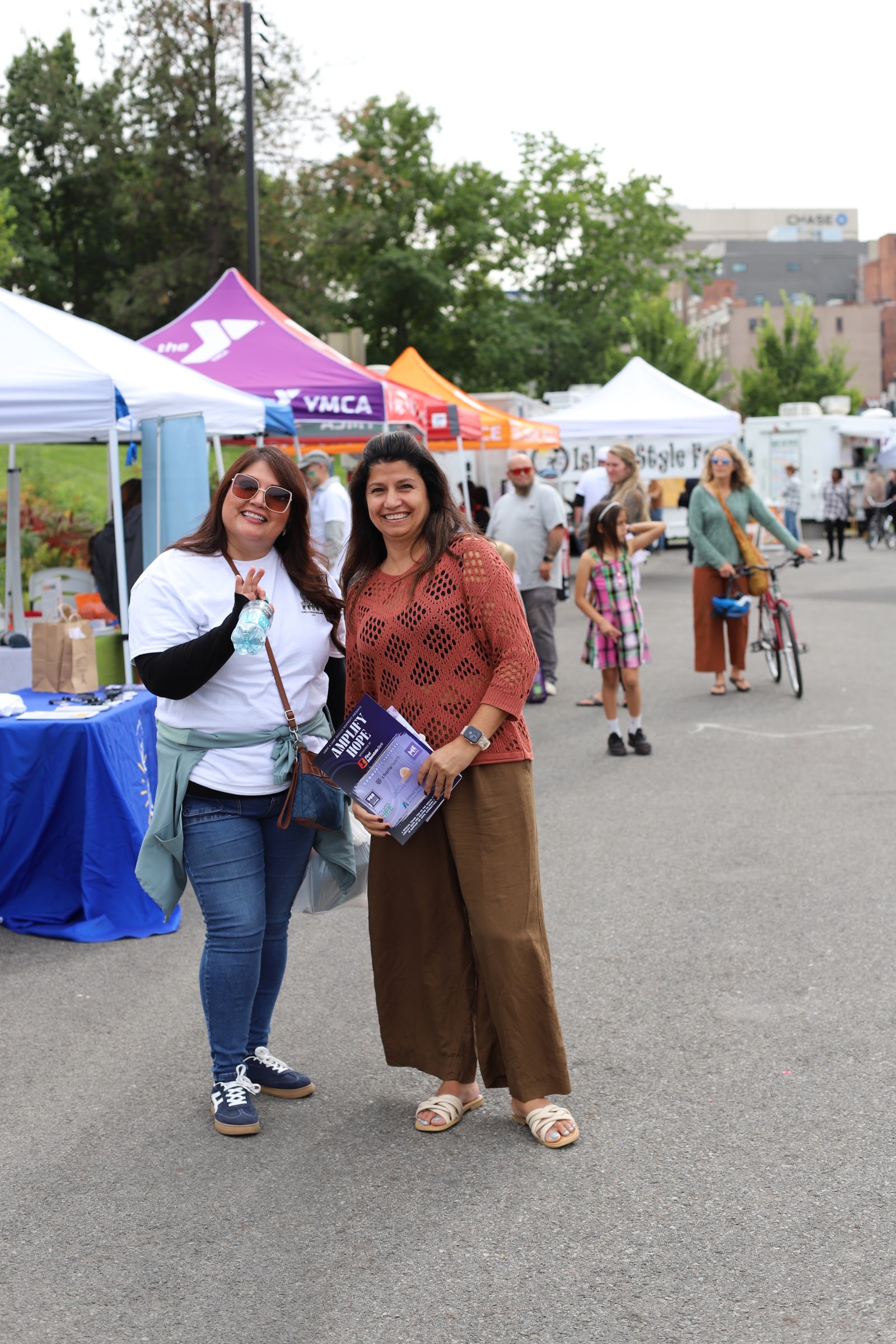 Two community members holding a shared outreach flyer, with multiple partner organizations' tents visible behind them at Hands Across Spokane.